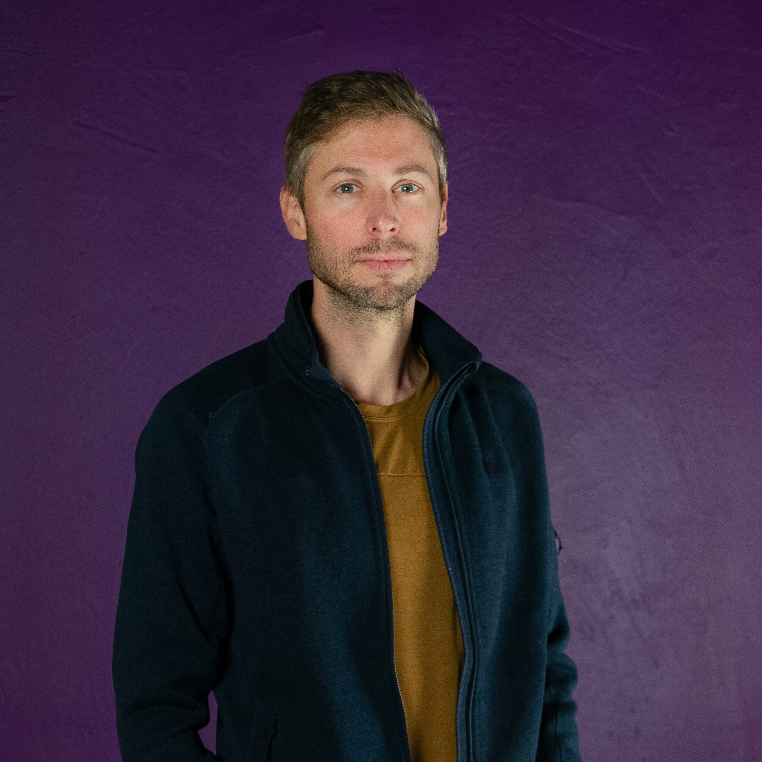 Become a member of Mautic 7 Photo of a white man with short, spiked light brown hair and a beard and moustache standing against a dark purple background. He's wearing a dark black jacket and a light brown coloured shirt.